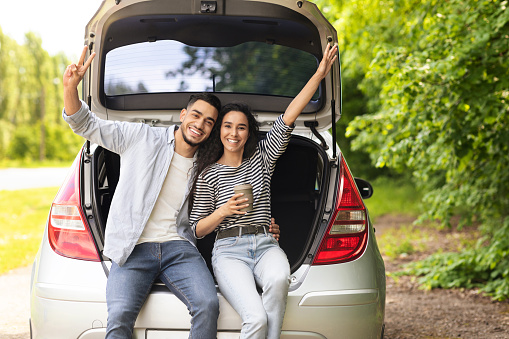 Emotional middle-eastern couple drinking coffee at country side, sitting on open auto trunk, embracing and gesturing, having coffee break while having car trip, enjoying vacation, copy space