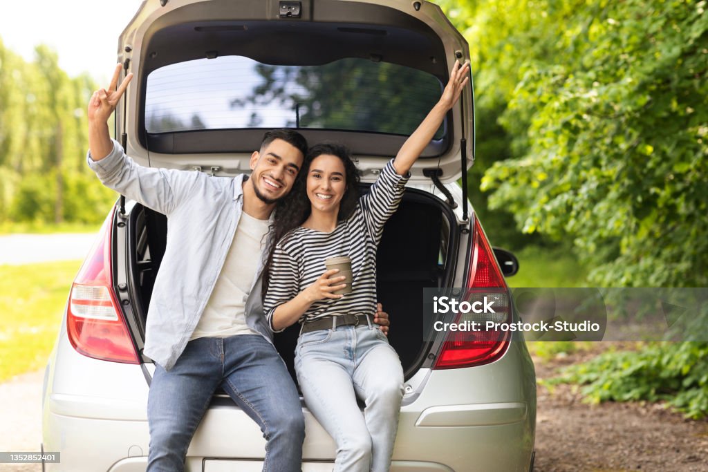 Emotional middle-eastern couple drinking coffee at country side Emotional middle-eastern couple drinking coffee at country side, sitting on open auto trunk, embracing and gesturing, having coffee break while having car trip, enjoying vacation, copy space Car Stock Photo