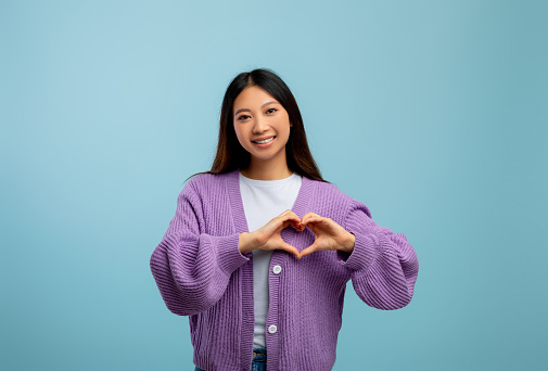 Kindness and love concept. Young asian lady making heart sign with fingers in front of her chest, smiling to camera over blue studio background