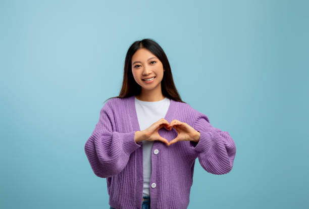 kindness and love concept. young asian lady making heart sign with fingers in front of her chest, blue background - doctor asian ethnicity chinese ethnicity young adult imagens e fotografias de stock