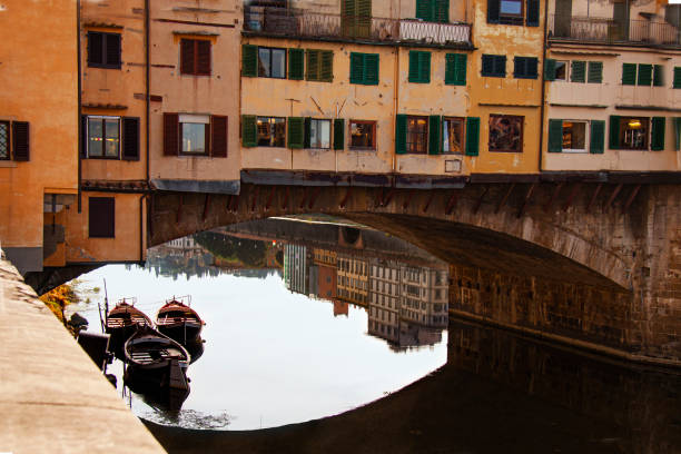 View of the Ponte Vecchio - Old Bridge - in Florence (Firenze) - by the river Arno with blue sky and reflections and boats in the water. Famous landmark in Tuscany, Italia.