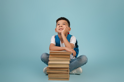 Portrait Of Dreamy Little Korean Schoolboy Leaning At Pile Of Books And Looking Up, Pensive Asian Male Child Wearing Backpack Sitting On Floor Over Blue Background In Studio, Copy Space
