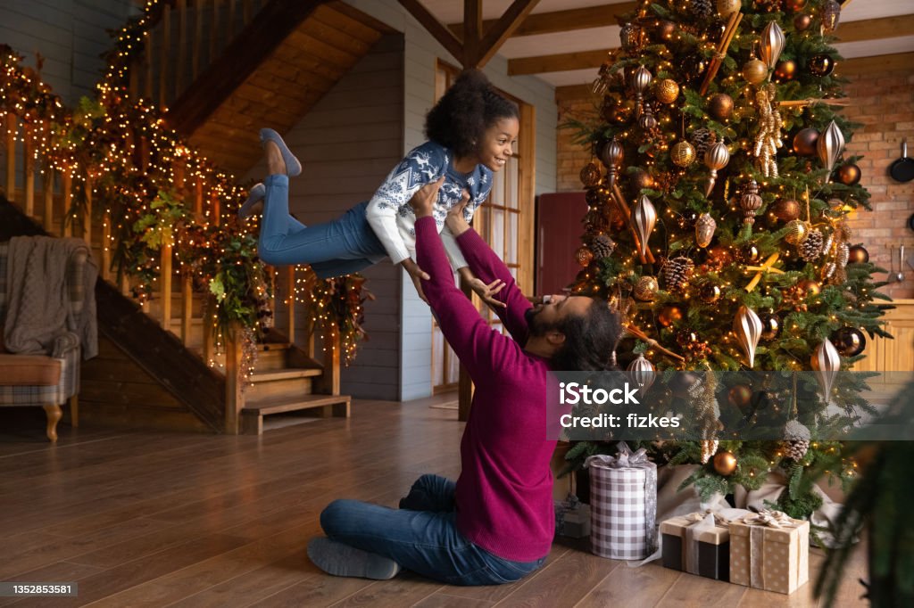 Playful biracial father have fun with small daughter at home Loving playful young African American father play with small daughter enjoy Christmas together. Smiling biracial dad engaged in funny game with little girl child on New Year winter holidays at home. Family Stock Photo