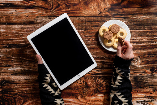 Woman's hands holding digital tablet and eating cookies on the wooden table