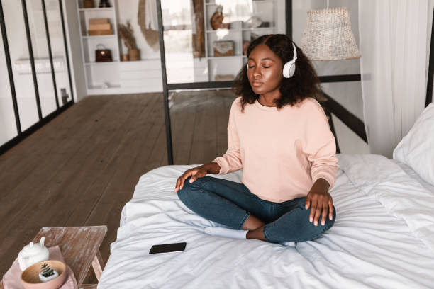 zrelaksowana afroamerykanka medytująca w słuchawkach w sypialni - lotus position audio zdjęcia i obrazy z banku zdjęć