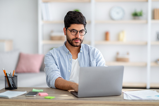 Trabajo remoto. Chico autónomo árabe concentrado que trabaja en una computadora portátil en la oficina en casa, sentado en el escritorio con computación photo