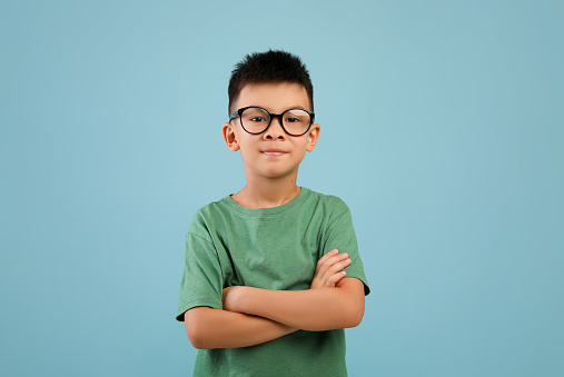Portrait of smiling little asian boy wearing eyeglasses standing with folded arms and looking at camera, confident nerdy korean male child posing over blue studio background, copy space