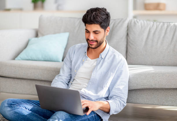 istock Young arab freelancer sitting with laptop on floor and leaning on sofa while working on computer at home interior 1352854123