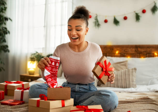 New Year celebration. Excited african american lady receiving xmas present and holding Christmas socks, sitting on bed New Year celebration. Excited african american lady receiving xmas present and holding Christmas socks, sitting on bed and unwrapping gift boxes unwrapping stock pictures, royalty-free photos & images