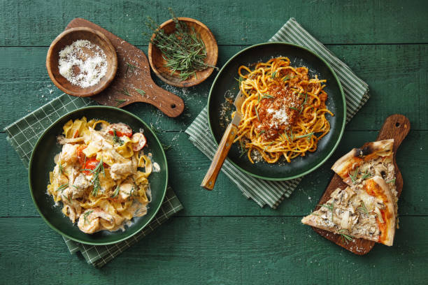 Italian Dishes for Family Dinner Classic spaghetti bolognese with parmesan cheese and herb. Italian fettuccine with prawns, salmon and herbs. Beef-mushroom pizza. Flat lay top-down composition on dark green background. NOODLES stock pictures, royalty-free photos & images
