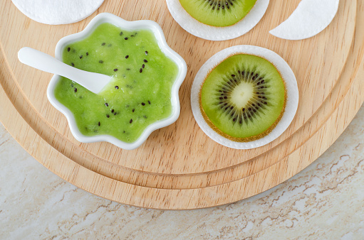 Fresh kiwi fruit puree in a small white bowl and cotton pads. Homemade face or eye mask, natural beauty treatment and spa recipe. Top view, copy space.