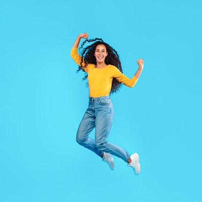 Joyful Arabic Woman Jumping Shaking Fists Over Blue Studio Background. Middle-Eastern Lady Posing In Mid-Air Gesturing Yes Celebrating Success And Having Fun. Joy Concept. Square