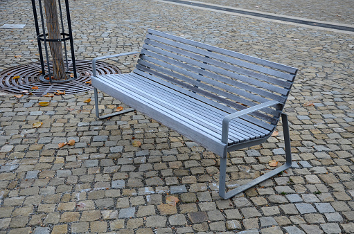 backrest, benches on a cobbled square with a sand surface of compacted gravel. ice icing. steel dark gray forniture for visitors. steel grilles in the shape of a circle allow trees to grow in the pavement,