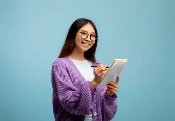 Photo of Checklist concept. Asian female student in glasses, taking notes in copybook and smiling to camera on blue background
