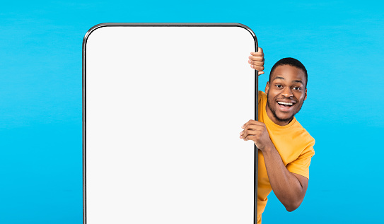Cheerful Black Guy Peeking Out Behind Big Smartphone With White Blank Screen, Excited African American Man Recommending New App Or Showing Copy Space For Mobile Website Design, Mockup Image