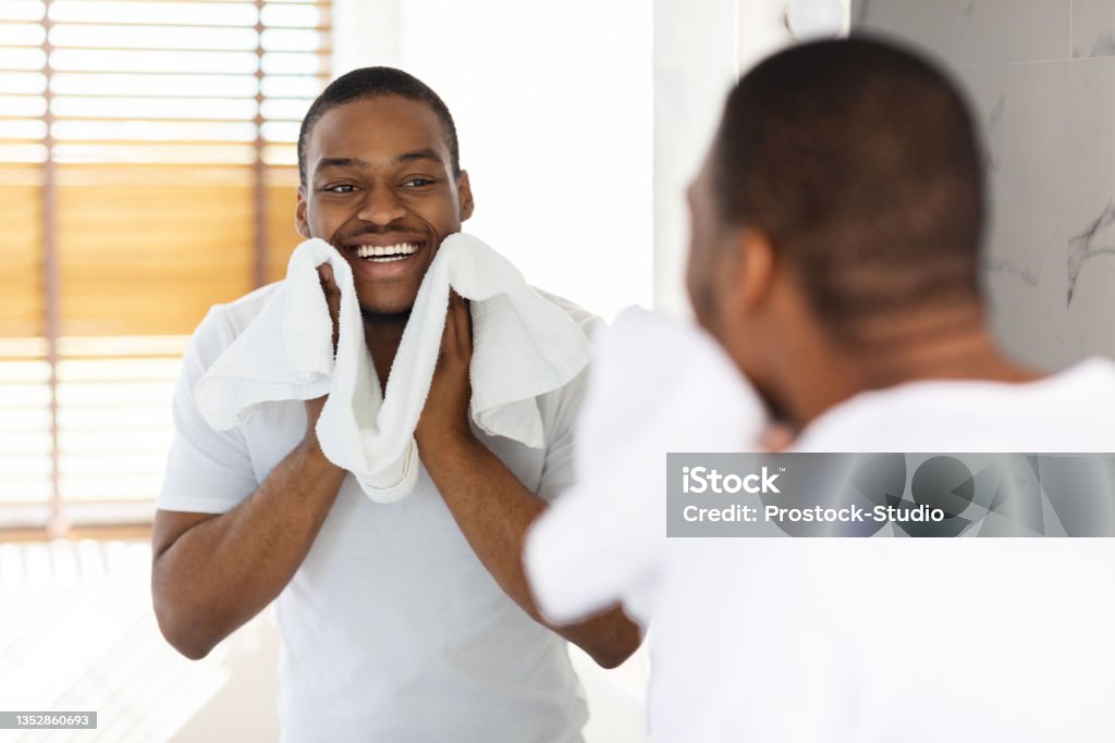 Aftershave Care Concept. Handsome Black Guy Wiping Face With Towel In Bathroom Aftershave Care Concept. Handsome Black Guy Wiping Face With Towel In Bathroom, Young African American Man Looking At Mirror And Smiling While Making Morning Hygiene, Selective Focus On Reflection Men Stock Photo