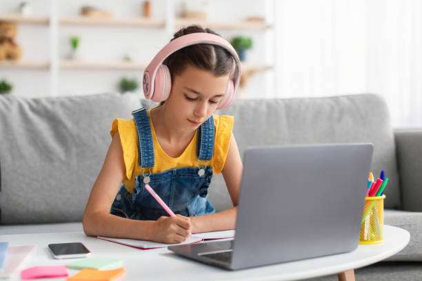 istock Schoolgirl in headset sitting at desk, writing in textbook 1352861857