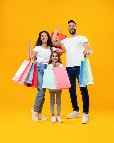 Middle-Eastern Family Holding Shopping Bags Smiling To Camera Standing On Yellow Studio Background. Parents And Daughter Posing With Colorful Paper Shoppers. Sales Season Advertisement. Vertical