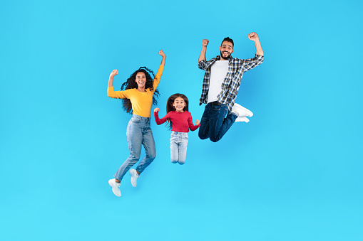 Joyful Arabic Family Jumping Shaking Fists Smiling To Camera Celebrating Success Over Blue Background. Middle-Eastern Parents And Daughter Posing In Mid-Air Rejoicing Victory In Studio. Full Length