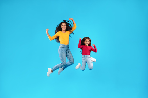 Happy Arabic Mother And Daughter Jumping Posing In Mid-Air Smiling To Camera Over Blue Background. Full Length Studio Shot Of Mom And Her Kid Celebrating Success Together And Having Fun