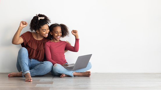 Emotional african american mom and kid gaming online together, using laptop, sitting on floor by white empty wall, raising hands up and smiling, playing video games, panorama with copy space