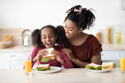 Loving black mother and daughter cuddling while having snack at kitchen, eating healthy sandwiches and drinking orange juice, happy mom embracing her hungry kid teen girl, copy space