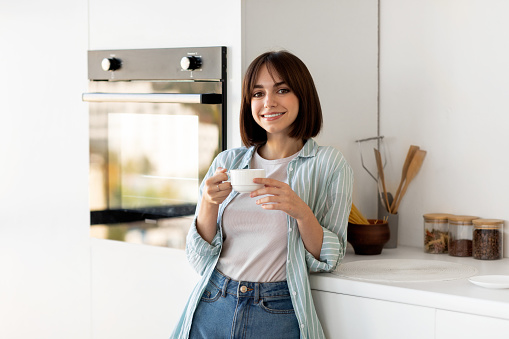 Home relax. Portrait of young lady drinking morning coffee in kitchen interior, holding cup with hot drink and smiling at camera, enjoying weekend, copy space