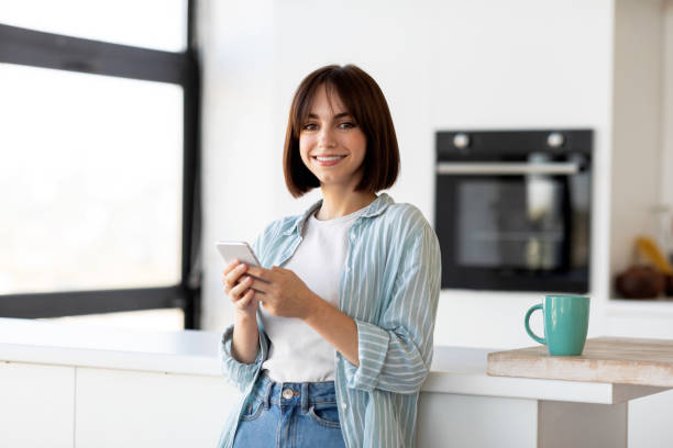 istock Smiling young lady using smartphone while enjoying coffee in kitchen at home, standing near table and smiling at camera 1352871315