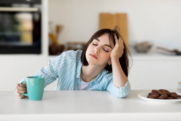 眠い若い女性がコーヒーを飲み、疲れを感じ、不眠症と睡眠障害に苦しみ、台所に座っている - だらしない女 ストックフォトと画像