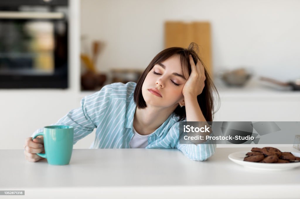 Sleepy young woman drinking coffee, feeling tired, suffering from insomnia and sleeping disorder, sitting in kitchen Sleepy young woman drinking coffee, feeling tired, suffering from insomnia and sleeping disorder. Sad female sitting in modern kitchen interior, empty space Tired Stock Photo