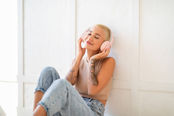 weekend relax. happy woman in wireless headphones listening music with closed eyes, sitting on floor over light wall - headphones women tattoo music imagens e fotografias de stock