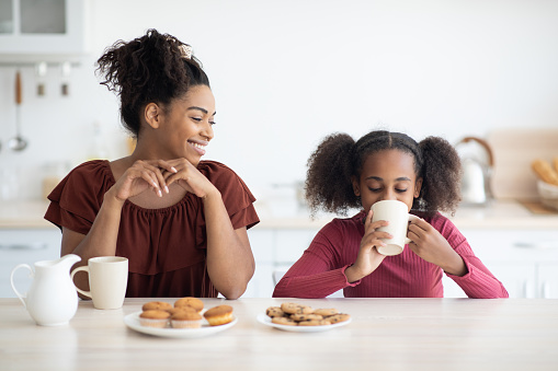 Beautiful black mother and daughter enjoying fresh pastry, sitting at kitchen table together, eating cupcakes and cookies, drinking tea and having conversation, spending weekend at home