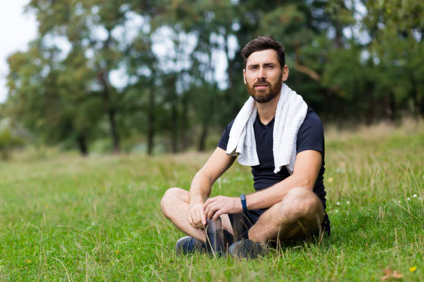 istock Exhausted after workout training or jogging athlete man sitting outdoors with a towel. Portrait sweaty fitness male in city park background forest and trees. Caucasian sportsman resting after running 1352874805