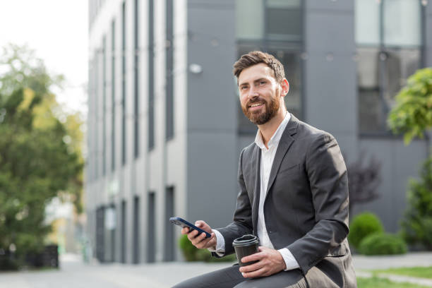 istock Handsome happy bearded businessman sitting on bench with cup coffee use mobile phone browse smartphone in hands reads good news and joyful smile. Outside a modern urban street background. City park 1352875101