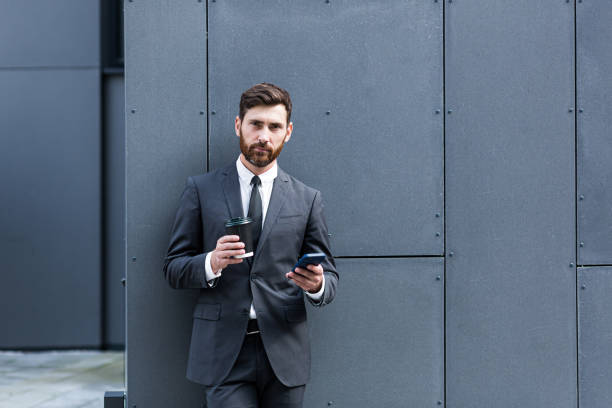istock Bearded businessman in formal suit on break using mobile phone use smartphone. business man standing outside on modern urban city street background with coffee cup in downtown outdoors. copy space 1352875165