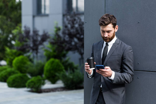 istock Bearded businessman in formal suit on break using mobile phone use smartphone. business man standing outside on modern urban city street background with coffee cup in downtown outdoors. copy space 1352875167