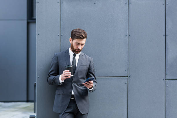 istock Caucasian bearded employee on urban background office building texting Messages, read good news mobile phone. Business man standing a formal suit messaging using Smartphone cellphone outdoors 1352875169