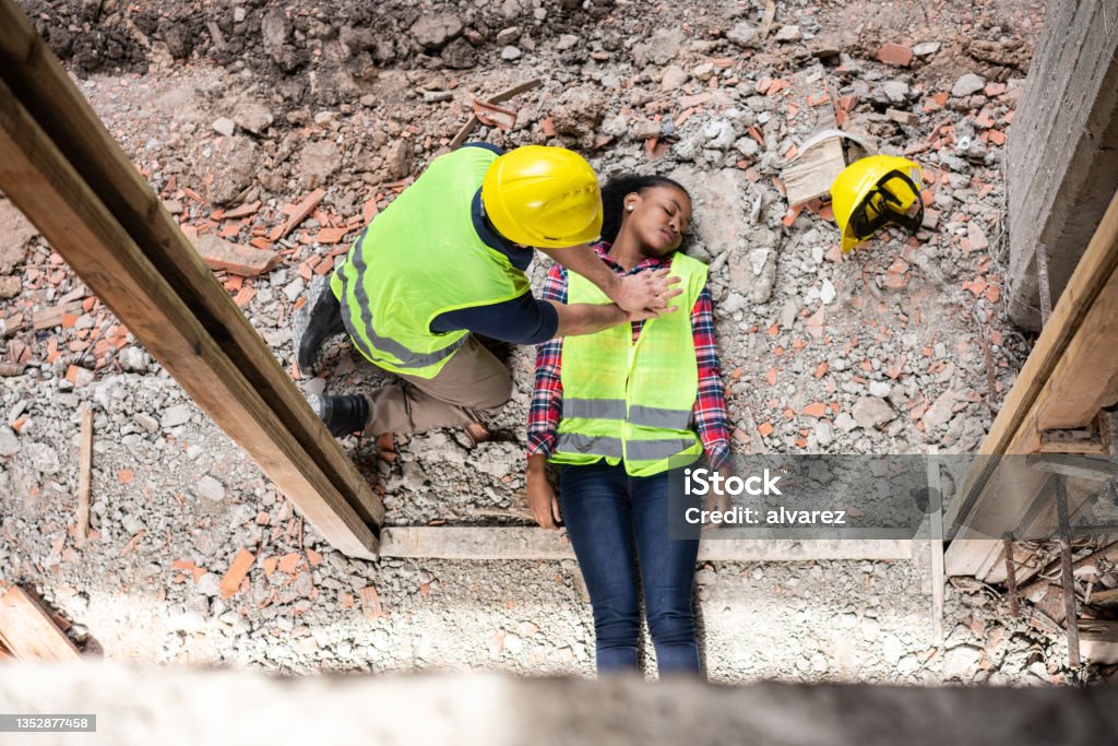 Construction worker giving first aid to a colleague lying on his back after an accident Accident of a woman worker at the construction site. Man performing cardiopulmonary resuscitation of a female worker. Top view of a construction worker giving first aid to a colleague lying on her back after an accident. CPR Stock Photo