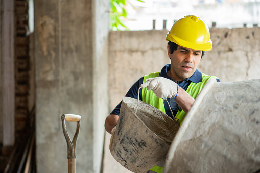 Male construction worker adding cement in the concrete mixer using bucket at a building site. Man worker manually loading the cement mixer machine at construction site.