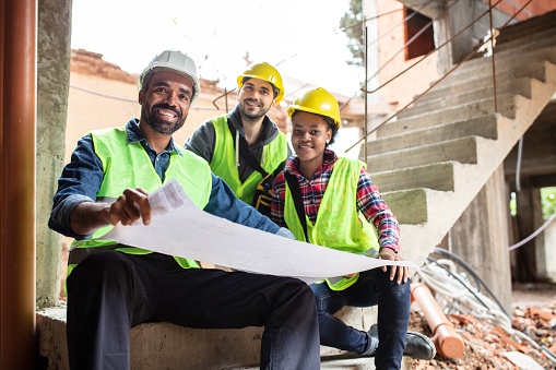 Portrait of group of three construction workers sitting together with building drawing. Multiracial builders looking at camera and smiling.
