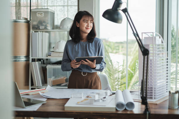 Portrait Asian woman Architect using tablet with blueprint and building model in office. stock photo