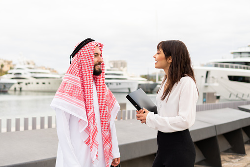 Two multiracial business partners standing in port and talking pleasantly after closing deal, young smiling european business woman discussing contract details with arab male client