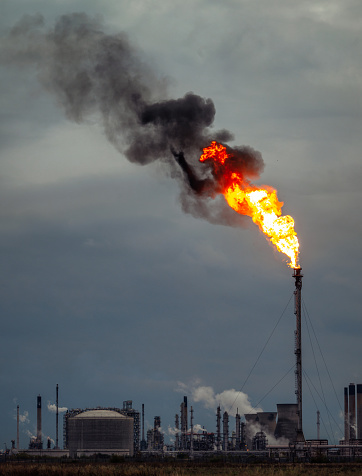 A large flame and black smoke polluting the air above a petrochemical plant at dusk.