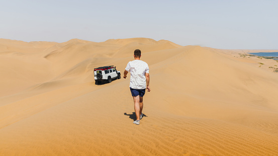Young man walking to her old truck car during the windy day at the sand dunes of Sandwich Harbour in Namib-Naukluft National park, Namibia, Souther Africa