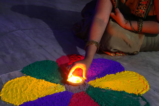 Stock photo showing a close-up view of a hand putting an oil filled diya in the centre of flower petal, gulal rangoli design, illuminated by flickering flame in the dark. Diwali festival of lights celebration. imagen de una persona irreconocible colocando diya encendida (lámpara de aceite) en el centro del diseño de gulal rangoli, patrón de pétalos de flores de pintura en polvo roja, púrpura, amarillo y verde, llama parpadeante en la oscuridad, celebración