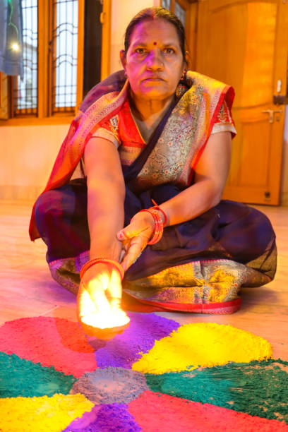 インドの女性がグラルランゴリデザインの中心に点灯したディヤ(オイルランプ)を置くイメージ、赤、紫、黄色、緑のパウダーペイント花びらのパターン、ちらつき炎、ディワリフェスティ� - bright brightly lit vibrant color burning ストックフォトと画像