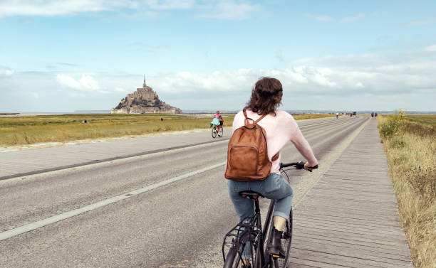 Young woman on bicycle wearing backpack with mont saint michel in background Young woman on bicycle wearing backpack with mont saint michel in background mont saint michel photos stock pictures, royalty-free photos & images