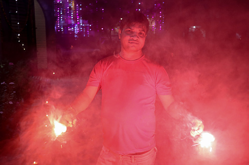 Stock photo of Hindu man dressed in casual clothing with house decorated with multicolour fairy lights. The man is holding lit sparklers to celebrate Diwali, the Indian traditional festival of lights.