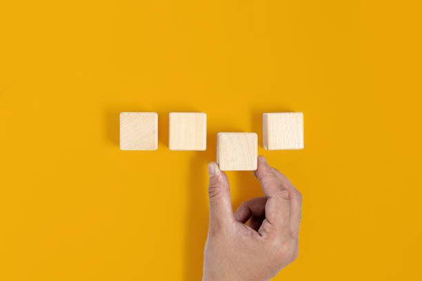istock A square wooden block is placed on a yellow background, the hand is picking up the third wooden block. Wood block concept, banner with copy space for text, poster, mockup template. 1352899847