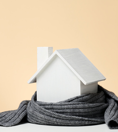 A white wooden miniature house wrapped in a gray knitted scarf. Building insulation concept, loans for repairs